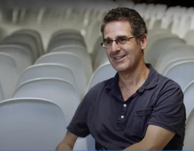 Mark Mazower, sorridente, sentado em frente a cadeiras brancas vazias. Sem texto na imagem. 