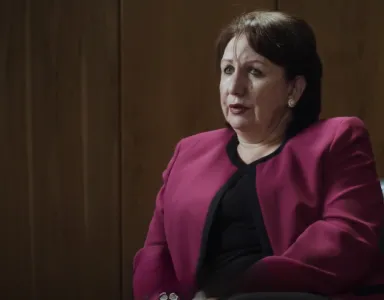 Mulher com blazer rosa sentado, expressão séria, num ambiente com paredes de madeira.