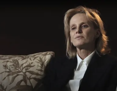 Imagem da mulher Siri Hustvedt, sentada num sofá com um padrão floral. Tem uma expressão  pensativa.