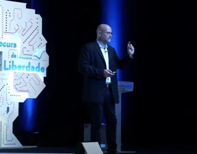 Yochai Benkler de pé num palco, gesticulando enquanto faz uma apresentação ao lado de um painel em forma de árvore com o texto "À procura da Liberdade".