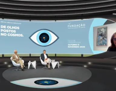 Carlos Fiolhais e outro homem sentados, num estúdio de televisão, a olhar para uma tela, na qual Ann Druyan está a falar.