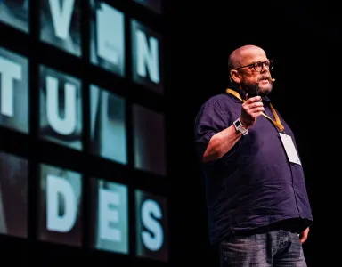 O sociólogo John Goodwin a falar em palco, a usar uma camisola escura e óculos. Texto visível: "juventudes".