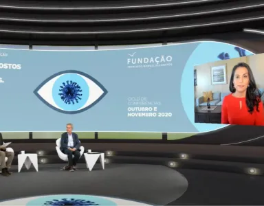 Dois apresentadores sentados, num estúdio, a olhar para uma tela na qual se vê Nahid Bhadelia a falar