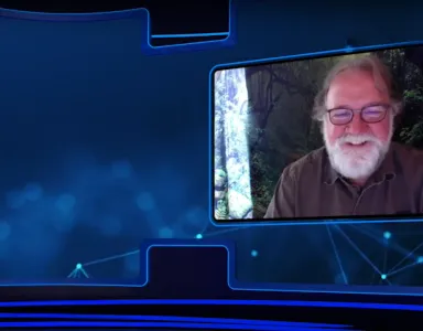 Sean B. Carroll, com barba e óculos, sorri durante uma videoconferência com fundo natural.