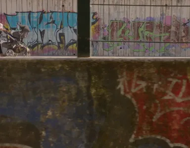 Mulher a passear com um bebé ao colo, enquanto empurra um carrinho, ao lado de uma parede com graffitis.