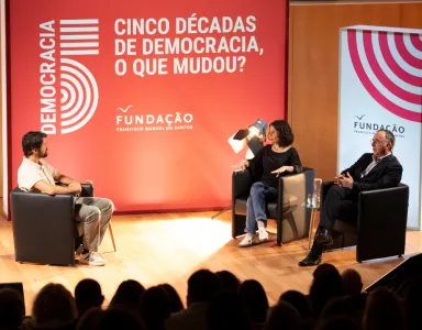 Evento sobre democracia com três pessoas em palco. Texto: "Cinco décadas de democracia, o que mudou?"
