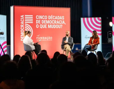 Tês pessoas num palco com público a assistir. Texto: "CINCO DÉCADAS DE DEMOCRACIA, O QUE MUDOU?"