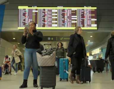 Mulher a olhar para o telemóvel em frente a um painel de chegadas e partidas do aeroporto de Lisboa.
