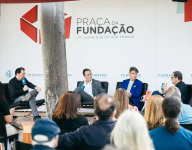 Imagem de um painel de discussão na Praça da Fundação, com quatro oradores e público a assistir.
