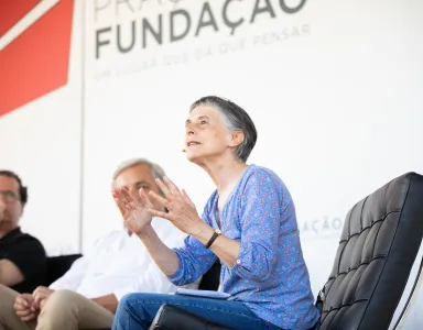 Mulher a falar e a gesticular, dois homens atrás, fundo com texto "FUNDAÇÃO".