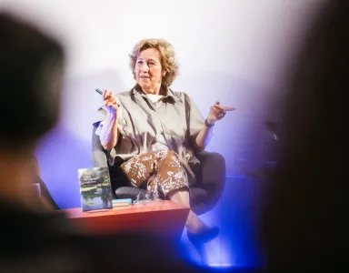 Mulher a falar em palco, sentada num sofá, com um livro na mesa à sua frente.