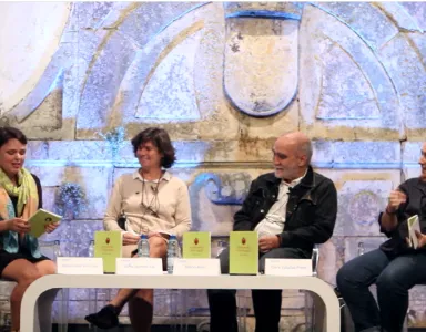 Evento literário com quatro pessoas sentadas, livros de capa verde na mesa.