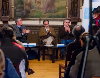 Evento com público a aplaudir, três oradores em destaque em frente a um quadro artístico.