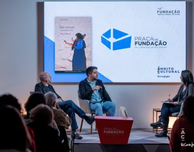 Uma plateia, de costas, a olhar para um palco, onde estão dois homens sentados a falar.