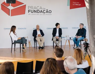 Quatro pessoas em palco, sentadas em cadeiras pretas, a falar com plateia a assistir. Logo e texto "Praça da Fundação".