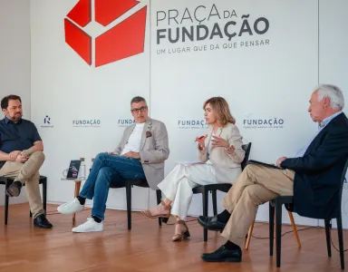 Quatro pessoas sentadas em cadeiras pretas, a debater entre si. Ao fundo, o texto "Praça da Fundação"