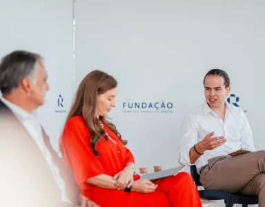 Três pessoas sentadas num palco. Dois homens de fato, uma senhora com um vestido vermelho. Ao fundo, o logotipo da Fundação.