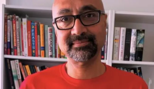 Imagem de um homem, o analista Bobby Gosh, vestido com uma t-shirt vermelha, com uma estante de livros atrás.