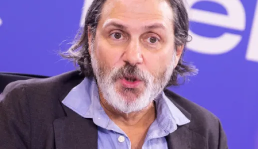 Retrato a cores de um homem com barba e cabelo grisalhos, vestido com uma camisa azul e um blazer preto, a falar para um microfone.
