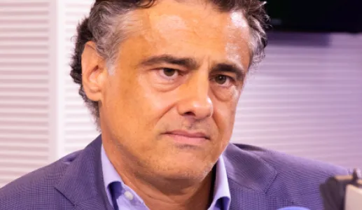 Um homem, vestido com um fato azul escuro e uma camisa azul clara, num estúdio de rádio, com um microfone à frente. Créditos: Fábio Oliveira.