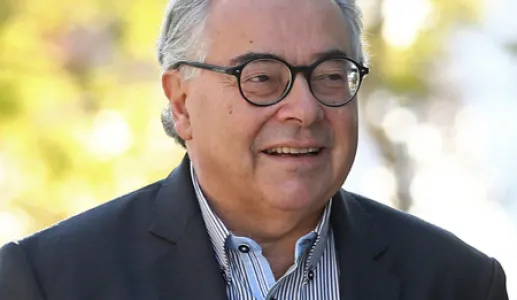 Retrato a cores de um homem de cabelo grisalho e óculos. Veste um blazer preto e uma camisa às riscas.
