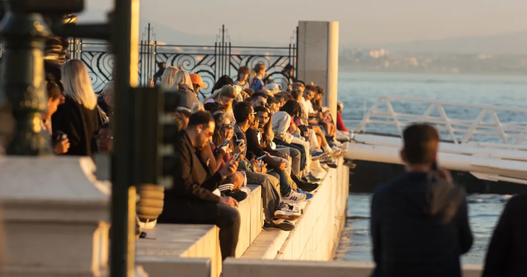 Pessoas de várias idades estão sentadas junto ao rio tejo, em Lisboa, ao entardecer. Crédito: Shutterstock