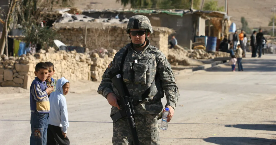 em grande plano um soldado norte-americano junto a uma estrada no Iraque, enquanto ao fundo várias crianças apontam para o militar. Crédito: Shutterstock