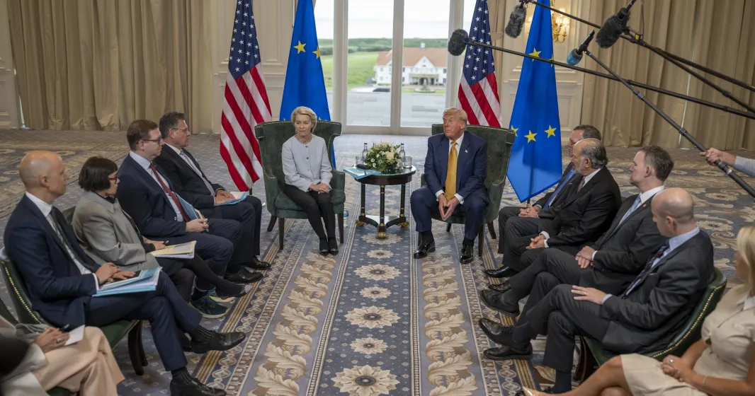 Ursula von der Leyen sentada ao lado de Donald Trump, com bandeiras da UE e dos EUA.