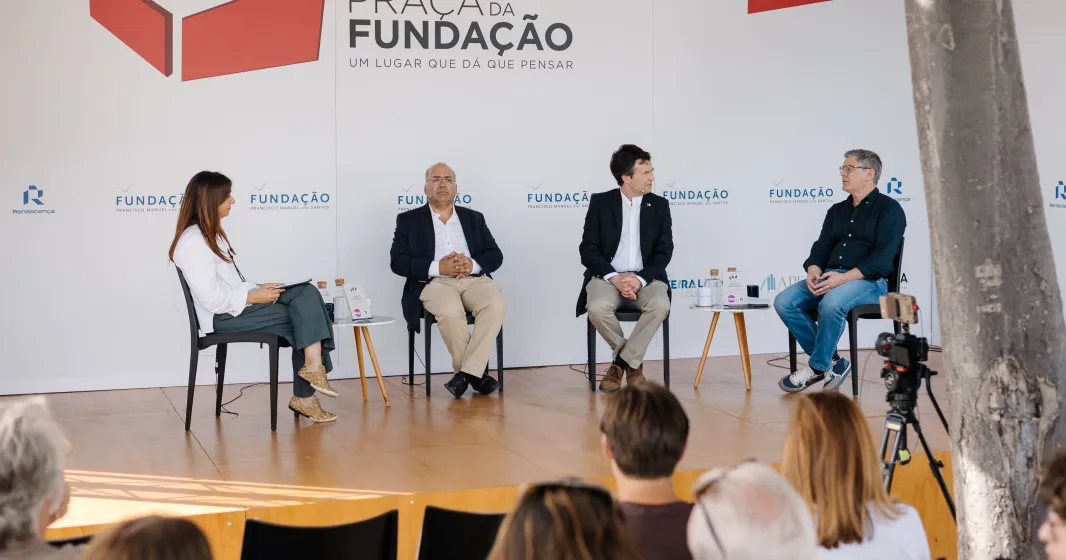 Quatro pessoas em palco, sentadas em cadeiras pretas, a falar com plateia a assistir. Logo e texto "Praça da Fundação".
