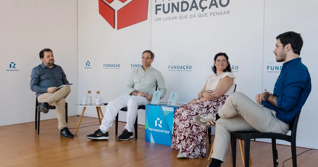 Quatro pessoas sentadas num palco, em cadeiras pretas, a apresentar um livro que está pousado em cima de uma mesa. Fundo com logotipos da Fundação.