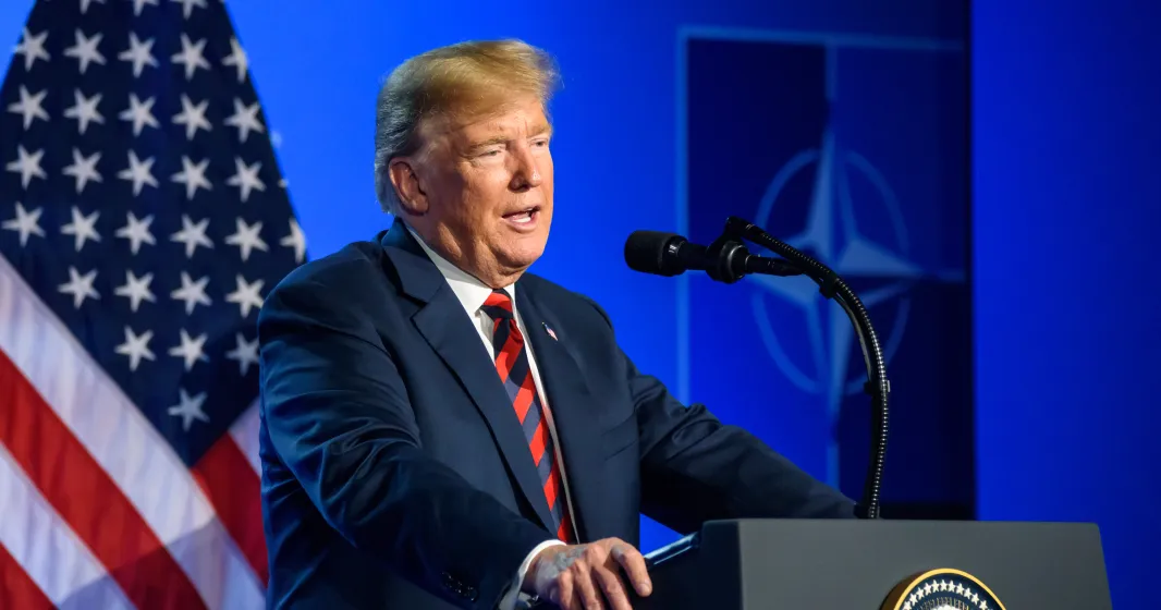 Donald Trump a falar, num púlpito. Fundo azul, com uma bandeira dos EUA e o símbolo da Nato. Créditos: Gints Ivuskans/shutterstock