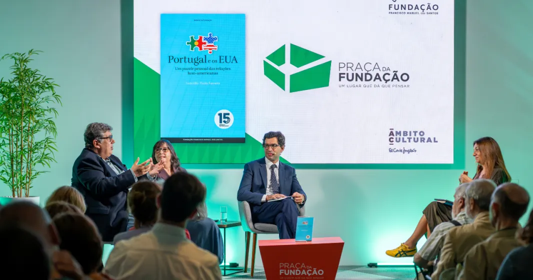 Imagem de quatro pessoas a conversar perante uma plateia,  sob um fundo verde.