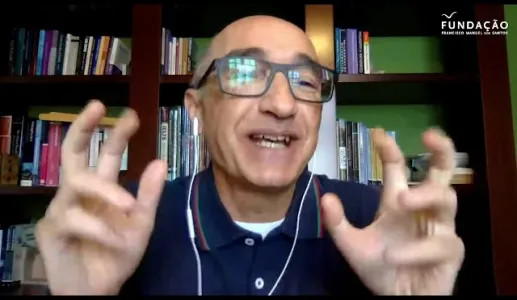 Homem com óculos, gesticulando em frente a uma estante de livros. Logotipo da Fundação.