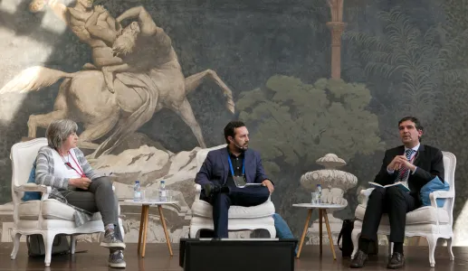 Três pessoas sentadas em cadeiras, a discutir em frente a um fundo artístico.