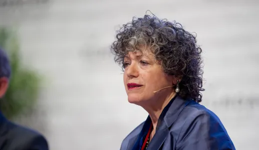 A professora Vivien A. Schmidt sentada, com um microfone sem fios, sobre um fundo branco. 