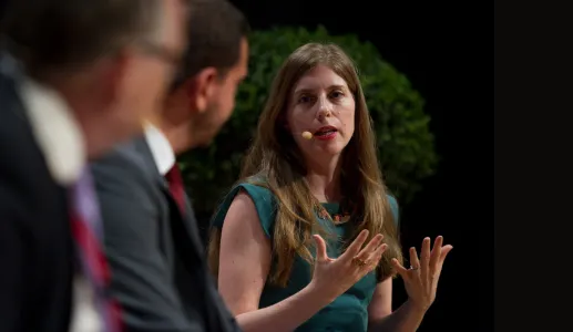 Mulher a falar num painel, a gesticular, e com um microfone na orelha. Fundo com plantas.