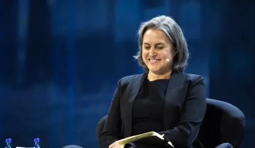 Mulher sorridente sentada em palco, segura um caderno. Luzes azuis ao fundo.