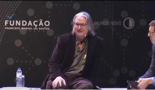 Bruce Sterling a falar, no palco da Casa da Música do Porto, sobre um fundo com o logo da Fundação Francisco Manuel dos Santos.