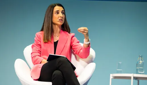 Mulher com blazer rosa fala, enquanto segura um caderno e gesticula. Fundo azul.