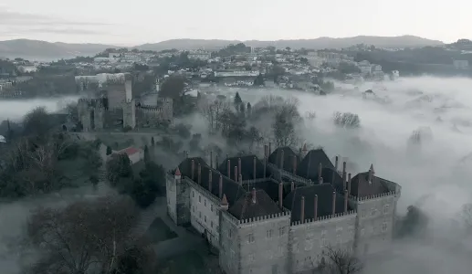 Castelo envolto em névoa, com vista para uma cidade nas colinas ao fundo.