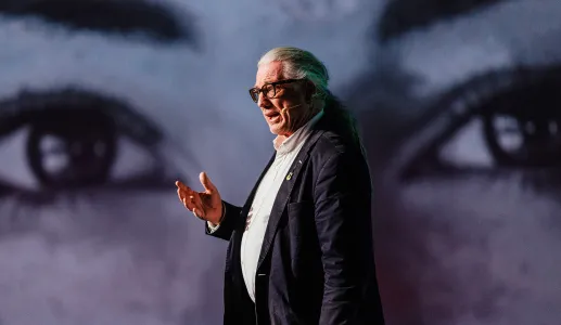 Howard Williamson, professor de Política Europeia, a falar de pé, gesticulando, sobre um fundo de dois olhos gigantes.