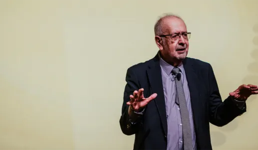 Um homem de óculos e fato, a gesticular durante uma apresentação. Fundo amarelo