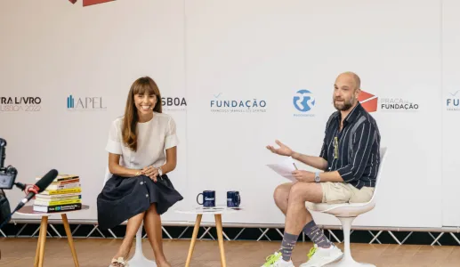Duas pessoas a conversar em palco. Duas mesas, uma com livros e outra com canecas. Fundo com vários logotipos.