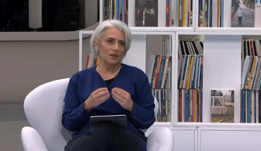 A jornalista Catarina Carvalho, sentada, a gesticular enquanto fala. Uma prateleira com livros ao fundo.