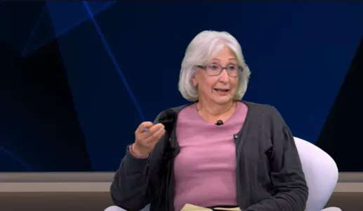 Mulher de cabelo grisalho, vestida com um casaco cinza e uma blusa rosa, a falar num estúdio de televisão.