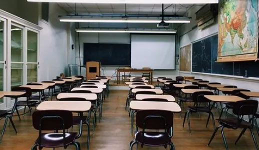Sala de aula vazia, com mesas e cadeiras dispostas, quadro negro e mapa na parede.