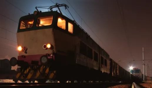 Locomotiva à noite com luzes acesas, transporte de vagões visível ao fundo.