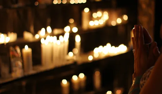 Mãos juntas em oração, diante de velas acesas, num espaço religioso.