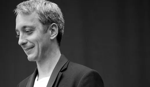 Homem sorridente com cabelo claro, vestido com um blazer preto. Fundo desfocado a preto e branco.