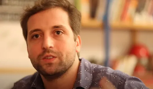 O humorista brasileiro Gregório Duvivier a falar, com uma estante de livros ao fundo.